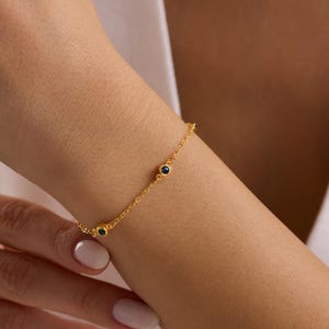 May include: A delicate gold bracelet with three small, dark blue gemstones. The bracelet is worn on a wrist, and the gold chain is visible against the skin. The bracelet is a simple, elegant piece of jewelry.