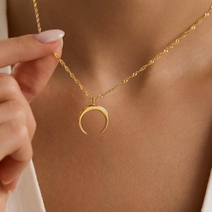 Può includere: Una collana con pendente a mezzaluna dorata. La collana presenta una catena delicata e un pendente curvo. La collana è tenuta da una persona che indossa un top bianco. Il pendente è un design semplice ed elegante.