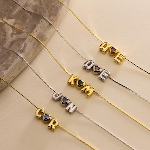 May include: A collection of delicate necklaces with letter pendants. Each necklace features a single letter pendant with a heart-shaped gemstone. The letters are in gold and silver tones, with colorful gemstones. The necklaces are displayed on a light beige surface.