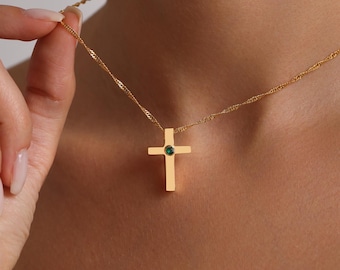 May include: Gold-colored cross pendant necklace with a small green gemstone at the center. The cross is rectangular with rounded edges. The necklace chain is a delicate, twisted design.