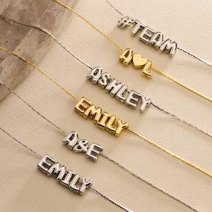 May include: A collection of delicate necklaces with personalized name pendants. The pendants are in silver and gold tones, spelling out names like "Ashley" and "Emily", and phrases like "#TEAM" and "A♥L". The necklaces are displayed on a neutral surface.