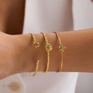 Peut inclure: Bracelets dorés avec des motifs floraux et étoilés. Les bracelets sont portés sur un poignet, mettant en valeur leurs détails complexes. L'un des bracelets présente une petite pierre bleue au centre d'une étoile.