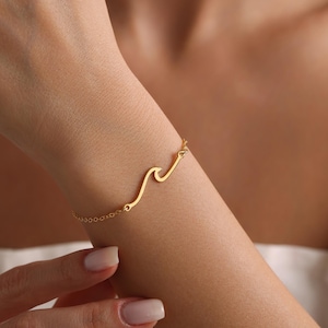 May include: A gold-colored bracelet featuring a wave design. The bracelet is worn on a wrist and is connected by a delicate chain. The wave design is a continuous line, creating a minimalist and elegant aesthetic. Jewelry.
