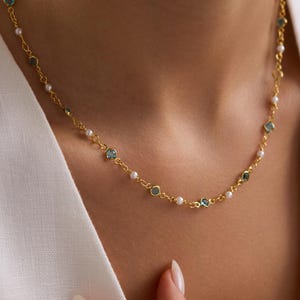Könnte beinhalten: Eine zarte goldene Halskette mit kleinen, runden, hellblauen Edelsteinen und winzigen weißen Perlen. Die Halskette ist an einer feinen Goldkette aufgereiht und wird vor einem weißen Hintergrund präsentiert.