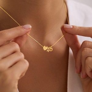 Peut inclure: Un délicat collier en or avec un charm en forme de cœur et le chiffre "45". Le collier est suspendu à une fine chaîne. Le cœur et les chiffres sont de couleur or. Le collier est tenu par une personne.