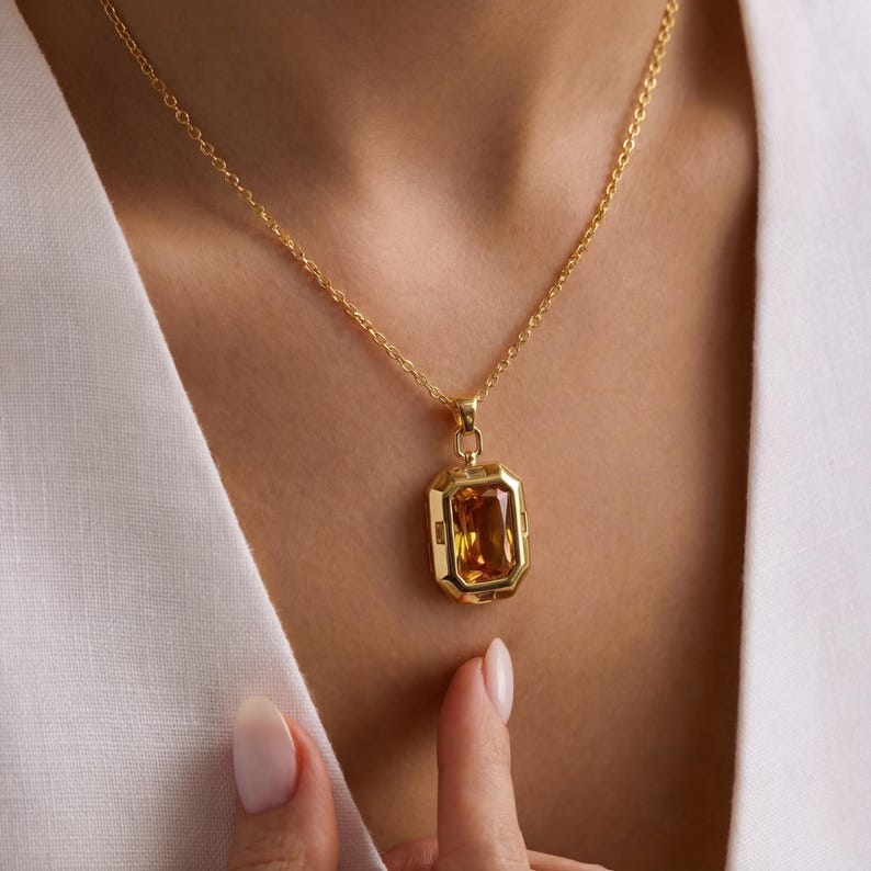 May include: Gold-toned necklace featuring a rectangular, amber-colored gemstone pendant. The pendant is set in a gold frame and hangs from a delicate gold chain. The necklace is displayed on a person wearing a white top.