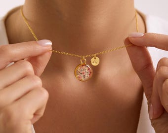 May include: A gold-colored necklace with a round pendant filled with small, colorful flowers. A small, round charm with the letter "A" hangs next to the pendant. The necklace is being held up by a person.