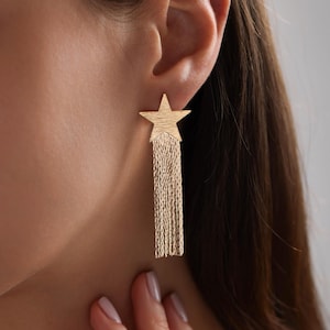 May include: Gold star-shaped earrings with a long chain tassel dangling from the bottom.
