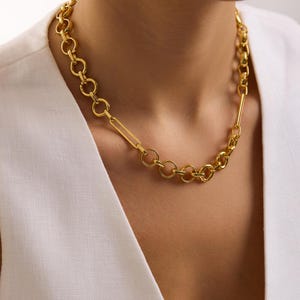 May include: A gold-colored necklace with a mixed chain design. The necklace features a combination of round and rectangular links, creating a bold and stylish accessory. The necklace is worn against a white top.