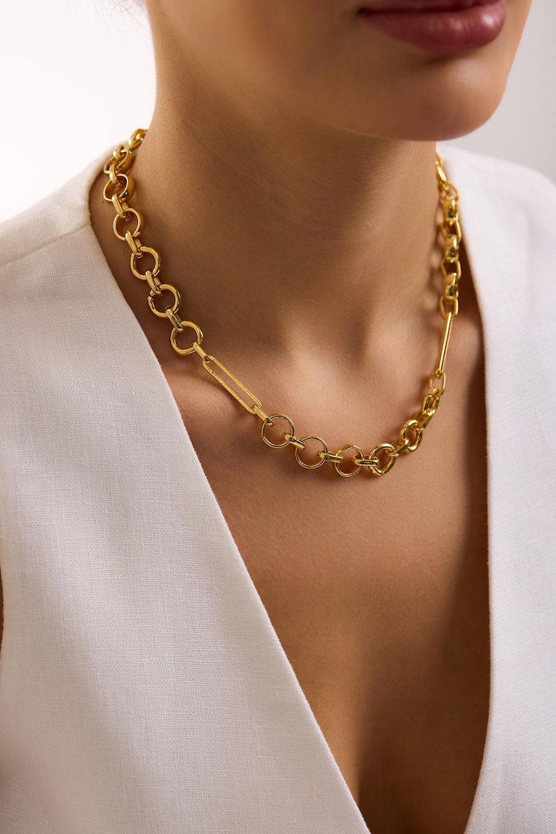 May include: A gold-colored necklace with a mixed chain design. The necklace features a combination of round and rectangular links, creating a bold and stylish accessory. The necklace is worn against a white top.
