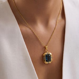 May include: A gold-colored necklace featuring a large, rectangular, dark blue gemstone pendant. The pendant is framed by a gold border with smaller blue gemstones. The necklace is worn against a white garment.