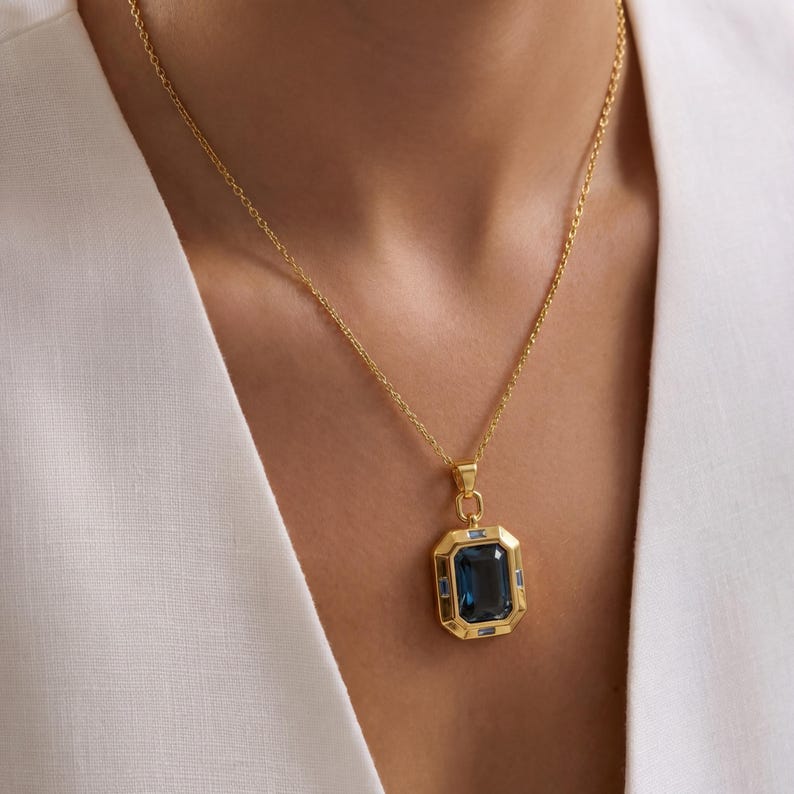 May include: A gold-colored necklace featuring a large, rectangular, dark blue gemstone pendant. The pendant is framed by a gold border with smaller blue gemstones. The necklace is worn against a white garment.
