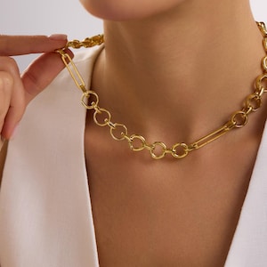 May include: Gold-toned chain necklace featuring a unique design. The necklace combines circular links with a large paperclip-style clasp. The jewelry is worn around the neck, showcasing its modern and stylish aesthetic.
