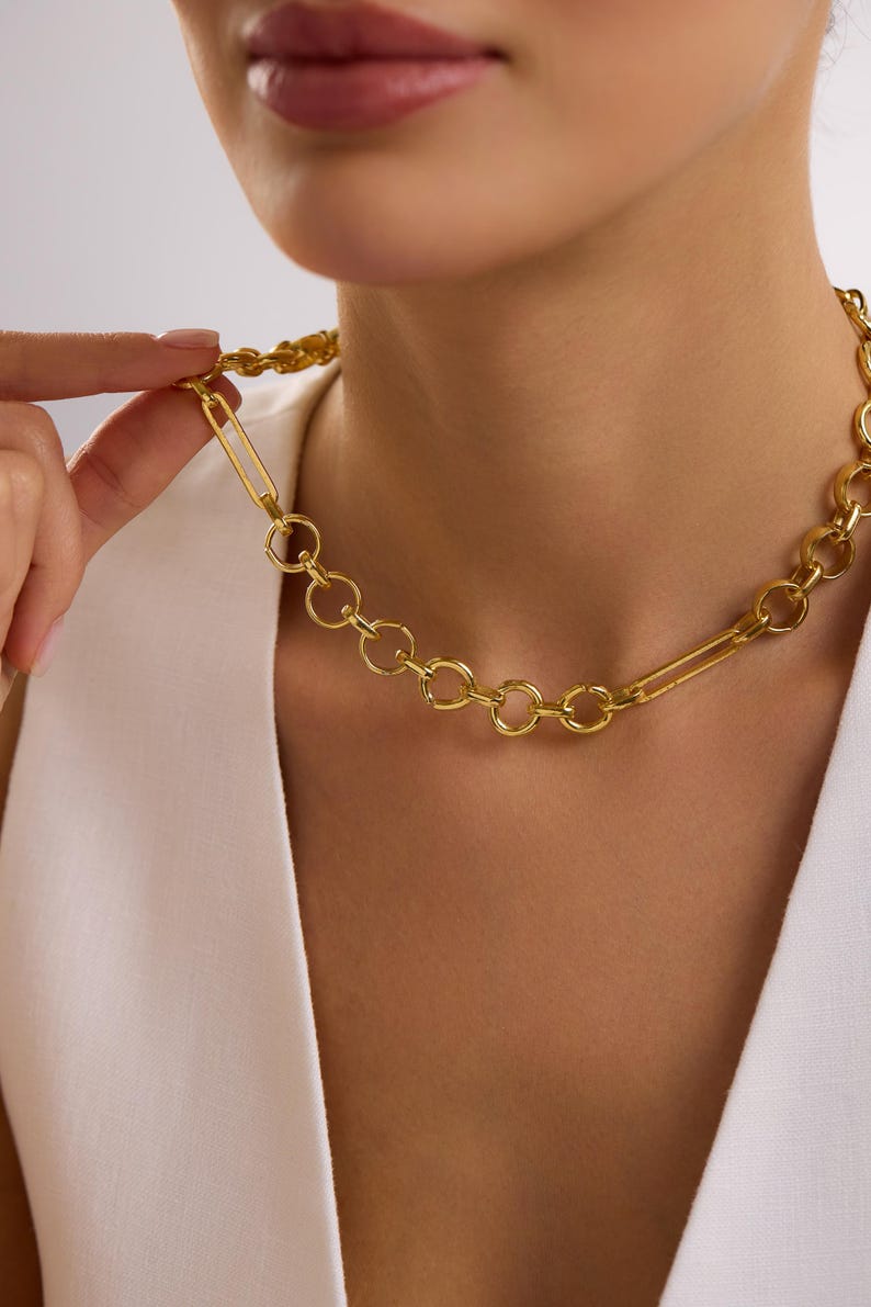 May include: Gold-toned chain necklace featuring a unique design. The necklace combines circular links with a large paperclip-style clasp. The jewelry is worn around the neck, showcasing its modern and stylish aesthetic.