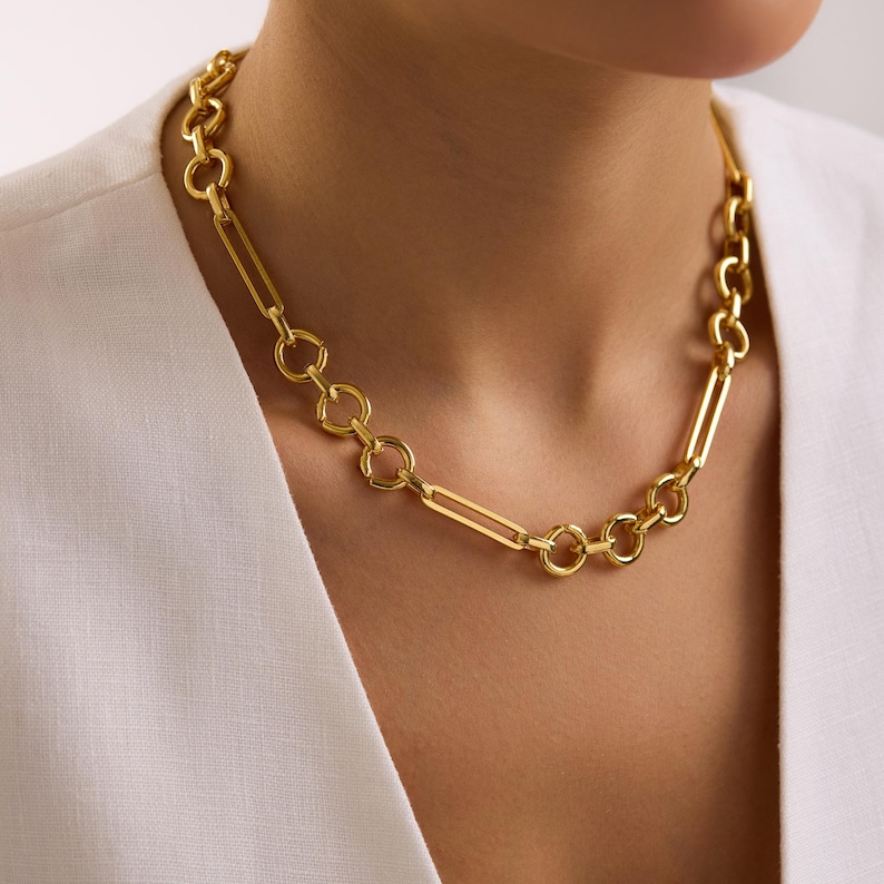 May include: A gold-colored chain necklace with a mix of elongated rectangular and circular links. The necklace is worn around the neck, and the background is a white shirt.