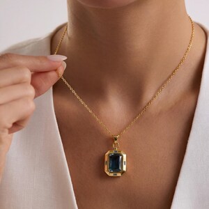 May include: A gold-colored necklace featuring a rectangular, dark blue gemstone pendant. The pendant is framed by a gold border and has smaller blue accent stones. The necklace is worn on a person with a white top.