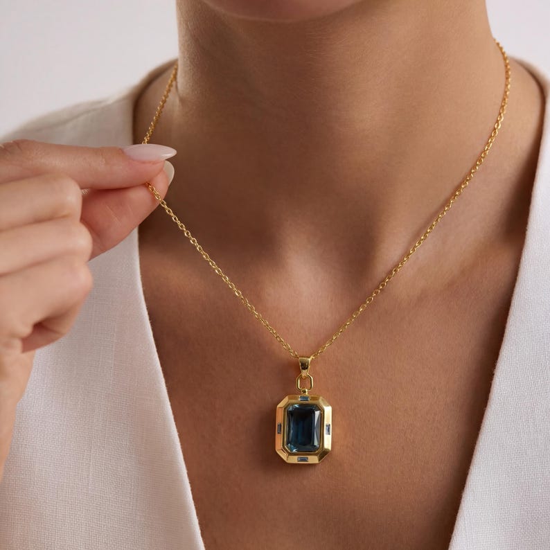 May include: A gold-colored necklace featuring a rectangular, dark blue gemstone pendant. The pendant is framed by a gold border and has smaller blue accent stones. The necklace is worn on a person with a white top.