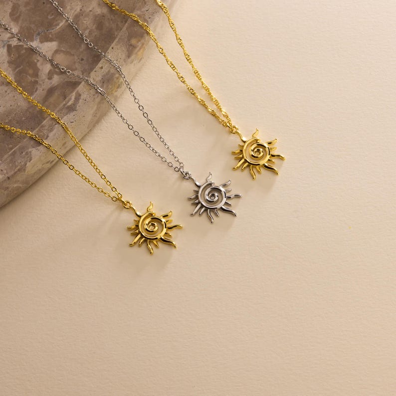 May include: Three necklaces with sun pendants. Two are gold-toned, and one is silver-toned. Each pendant features a spiral center with radiating rays. The chains are delicate and hang at varying lengths, creating a layered effect.