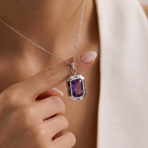 May include: Silver necklace with a rectangular amethyst pendant. The pendant has a large, central purple gemstone surrounded by smaller purple stones and a silver frame. The chain is delicate and the overall design is elegant.