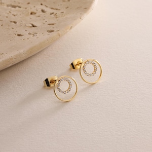 May include: A pair of gold-toned earrings with a circle design. The circle is outlined with a thin gold wire and has a row of small clear stones around the edge.