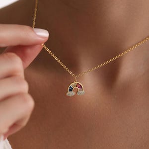 May include: A gold chain necklace with a rainbow pendant. The rainbow is made of colorful stones and is surrounded by small clear stones. The pendant is shaped like a rainbow with clouds on either side.