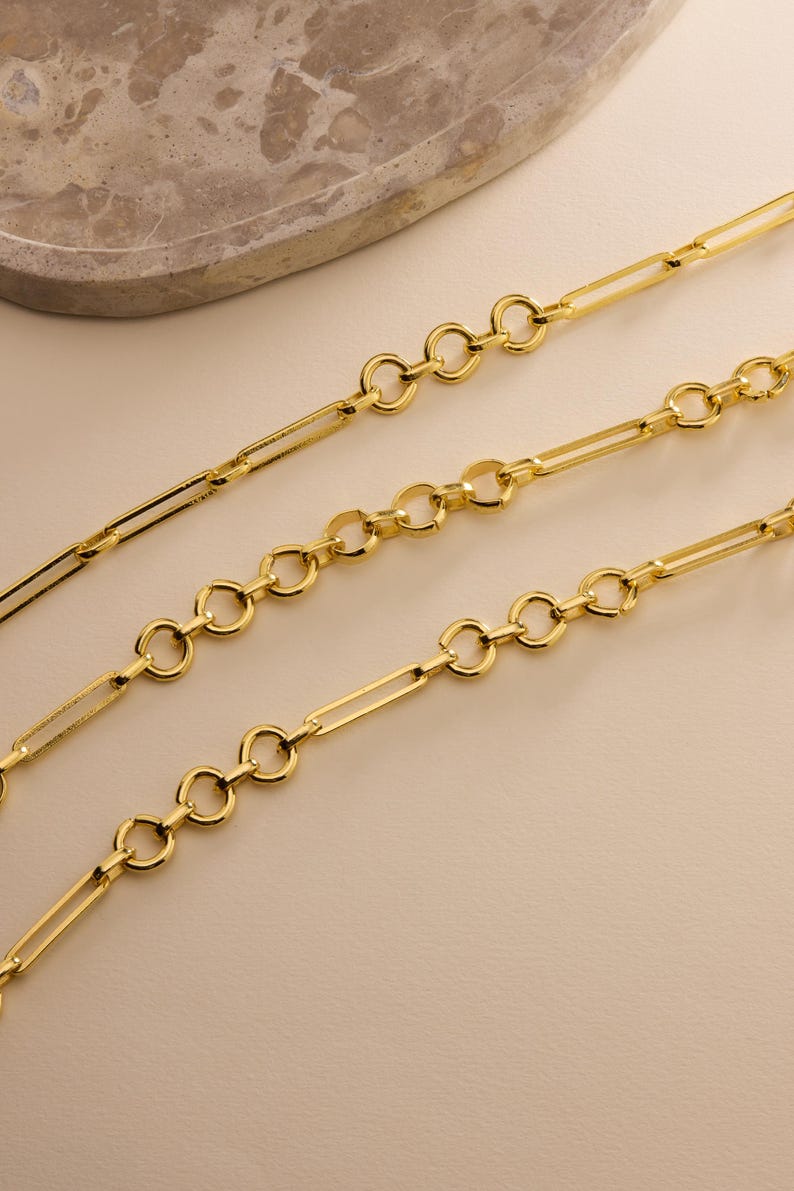 May include: Three gold-colored chain necklaces with alternating oval and rectangular links. The chains are displayed on a light beige surface, with a marble-like accent in the background. The jewelry is likely for fashion or personal adornment.