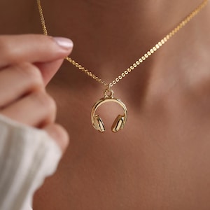 Puede incluir: Un collar de cadena dorada con un colgante de auriculares dorados. El collar lo lleva una persona.