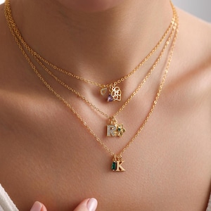 Puede incluir: Un collar de oro de varias capas con tres colgantes: un corazón con una piedra morada, una letra 'R' con una flor y una letra 'K' con una piedra verde.