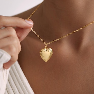 Puede incluir: Un collar de oro con un dije en forma de corazón y una cadena delicada. El dije tiene un diseño adornado y está cerrado.