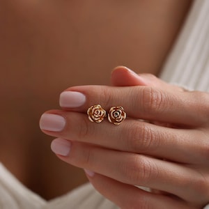 May include: A pair of gold-colored rose stud earrings. The earrings are small, with detailed petals, and are held in a hand with manicured nails. The background is blurred, focusing attention on the jewelry.