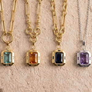 May include: Four necklaces with gemstone pendants. The first three have gold chains and pendants with rectangular gemstones in teal, orange, and navy blue. The fourth necklace has a silver chain and a purple gemstone pendant.