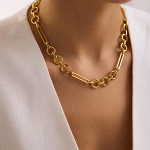 May include: A gold-colored necklace with a mix of circular and rectangular links. The necklace is worn around the neck, and the links are connected to each other. The necklace is a bright gold color and appears to be made of metal.