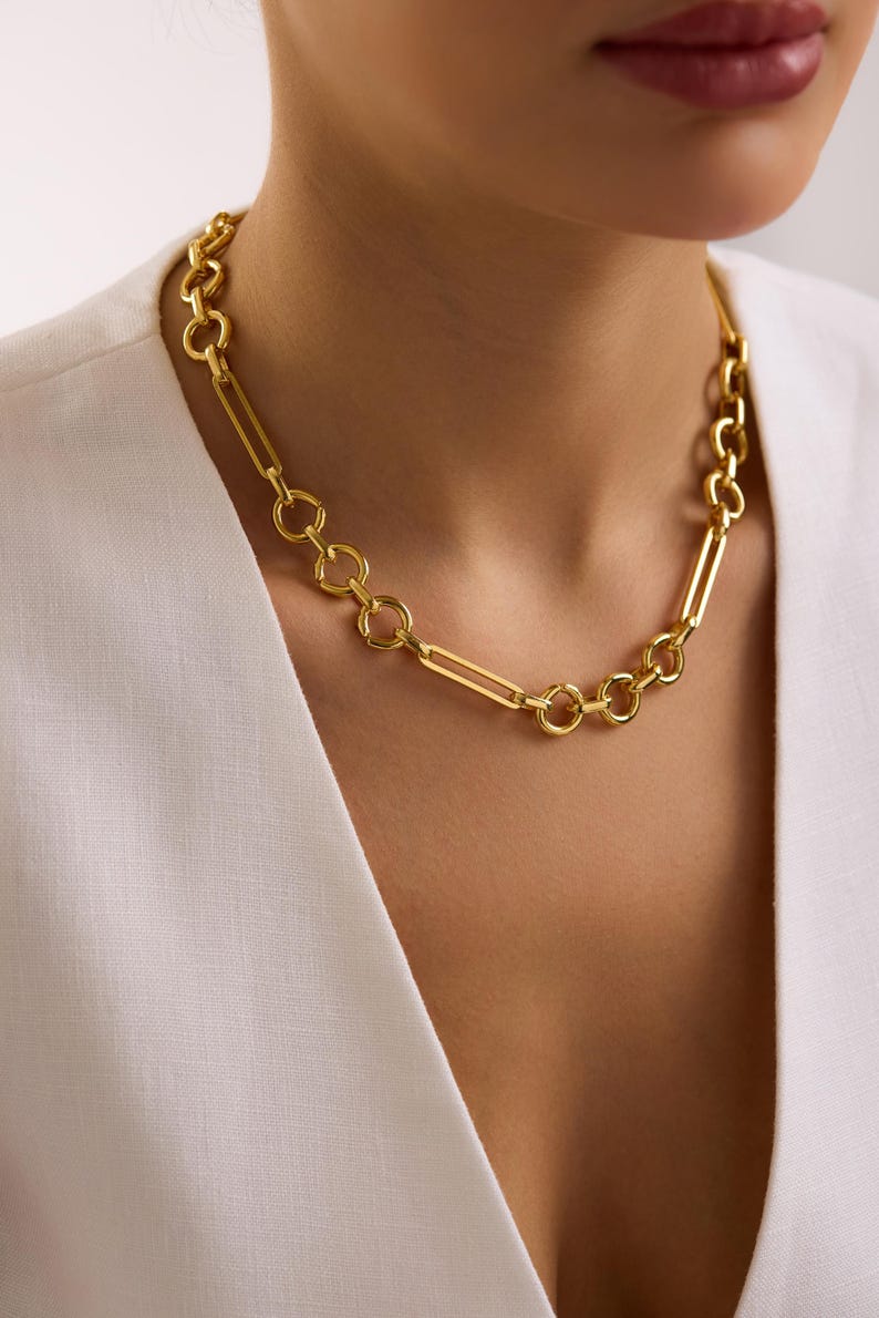May include: A gold-colored necklace with a mix of circular and rectangular links. The necklace is worn around the neck, and the links are connected to each other. The necklace is a bright gold color and appears to be made of metal.