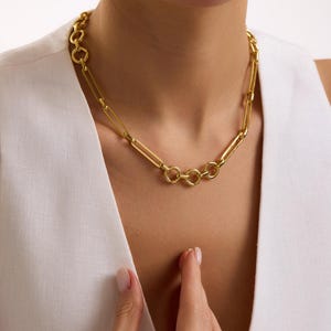 May include: A gold-colored necklace with a chain-link design. The necklace features a series of elongated rectangular links interspersed with circular links. The jewelry is worn around the neck.