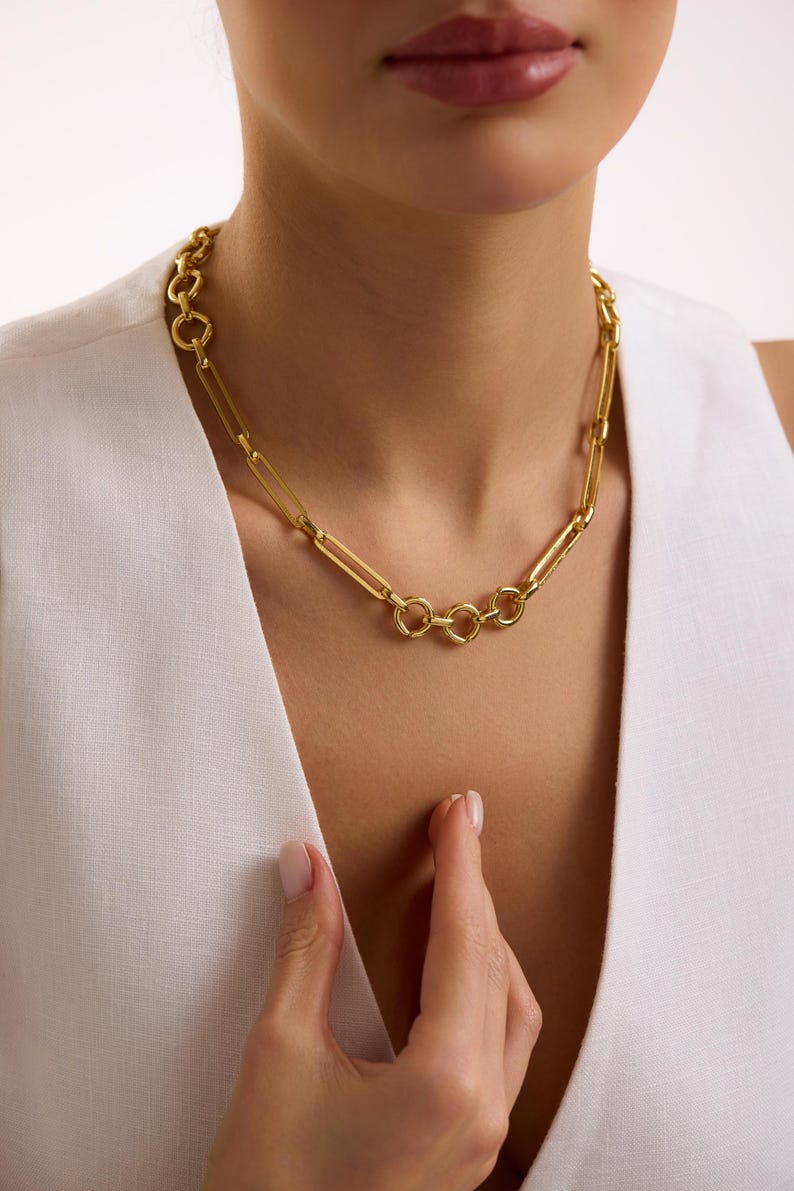 May include: A gold-colored necklace with a chain-link design. The necklace features a series of elongated rectangular links interspersed with circular links. The jewelry is worn around the neck.