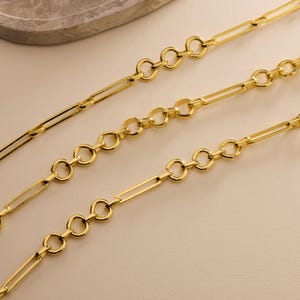 May include: Three gold-colored chain necklaces with different link designs. One chain features elongated rectangular links, while the others have a combination of circular and rectangular links. The chains are displayed on a neutral background.