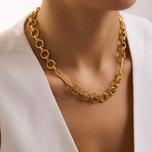 May include: A gold-toned necklace with a mixed chain design. The necklace features a combination of circular links, a rectangular link, and a thicker chain section. The jewelry is worn against a white top.