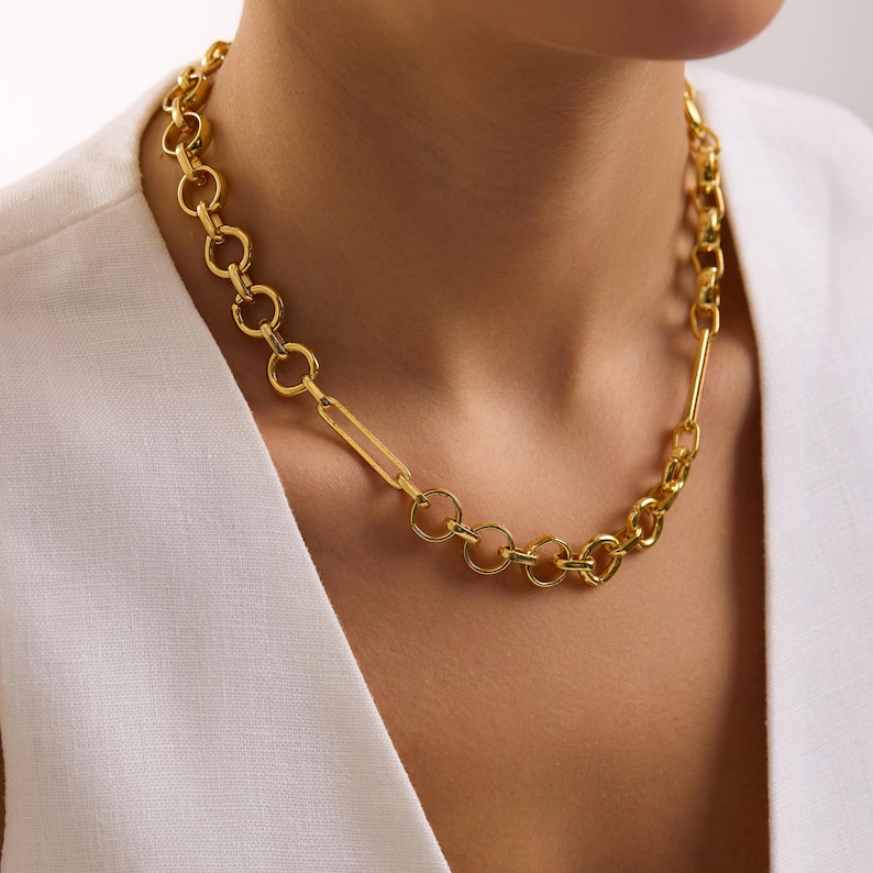 May include: A gold-toned necklace with a mixed chain design. The necklace features a combination of circular links, a rectangular link, and a thicker chain section. The jewelry is worn against a white top.