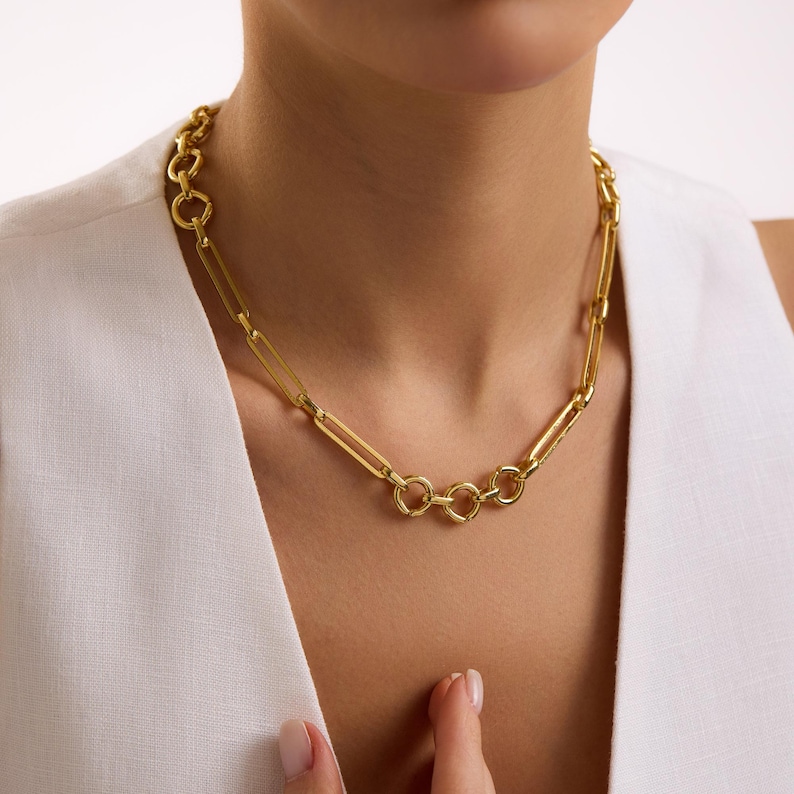 May include: A gold-colored chain necklace with a mix of elongated rectangular and circular links. The necklace is worn around the neck and is a bright, shiny gold color. The necklace is a statement piece.