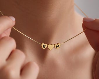 May include: A gold chain necklace with three heart-shaped charms. Each charm has a letter engraved on it, spelling out "E M".