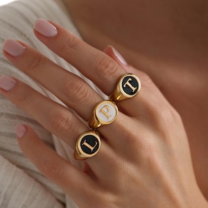 Puede incluir: Tres anillos de sello dorados con caras de esmalte negro y blanco, cada uno con una letra diferente: L, P y T. Los anillos son de forma ovalada y tienen un acabado pulido. Las letras están en una fuente serif.