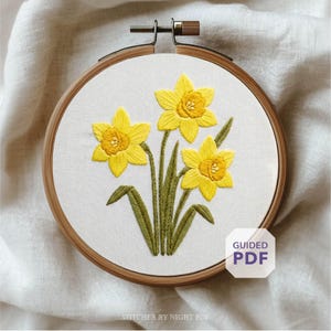May include: A hand-stitched embroidery hoop featuring three yellow daffodil flowers with green stems and leaves. The embroidery is set against a white fabric background, framed by a wooden hoop. The text "GUIDED PDF" is in the lower right corner.
