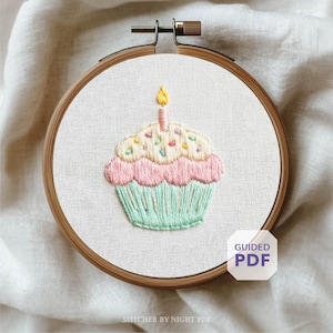 May include: Embroidered cupcake design in a wooden hoop. The cupcake features pink frosting, a light green base, and a yellow candle. The design includes colorful sprinkles. The text "Guided PDF" is visible.
