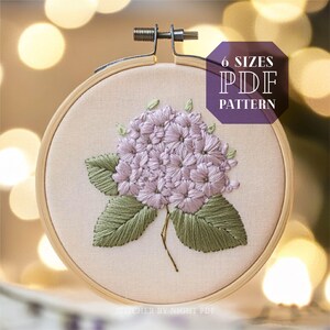 May include: A close-up of a hoop with a delicate embroidery design of a hydrangea flower with green leaves. The hoop is on a light background with a blurred out bokeh effect. The text "6 SIZES PDF PATTERN" is in the upper right corner of the image.
