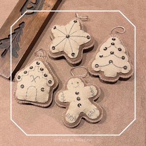 May include: Four Christmas ornaments in various shapes: a star, a Christmas tree, a gingerbread house, and a gingerbread man. Each ornament is made of off-white felt with silver stitching and embellishments, and a tan border. They have loops for hanging.