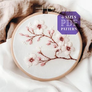 Puede incluir: Un aro de bordado blanco con un delicado diseño de bordado de una rama con flores rosas y blancas. El texto "6 SIZES PDF PATTERN" está en un cuadro morado en el lado derecho de la imagen.