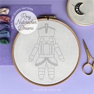 May include: An embroidery hoop with a line drawing of a nutcracker, featuring a crown and mustache. The text "Rosy Nutcracker Dreams" is above the design. The image includes embroidery floss, a needle minder, and gold scissors.