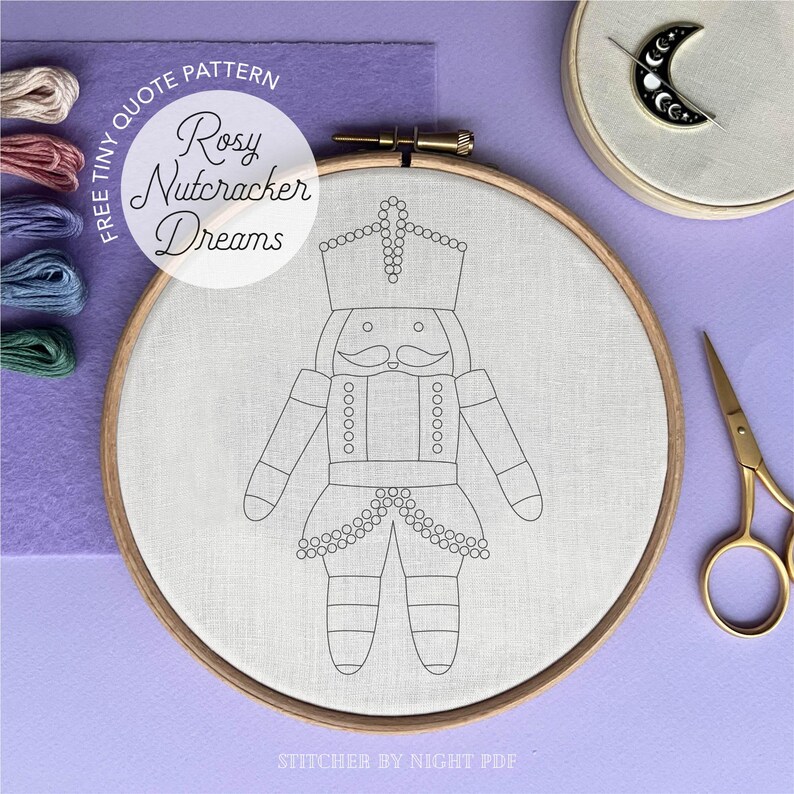 May include: An embroidery hoop with a line drawing of a nutcracker, featuring a crown and mustache. The text "Rosy Nutcracker Dreams" is above the design. The image includes embroidery floss, a needle minder, and gold scissors.