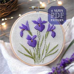 May include: Embroidered iris flower design in a wooden embroidery hoop. The design features purple irises with green leaves and stems. The hoop is on a white fabric background. 6 Sizes PDF Pattern.