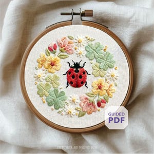 May include: Embroidered hoop art showing a red ladybird with black spots, encircled by a wreath of colourful flowers and green leaves. The embroidery is set in a wooden hoop. The text "GUIDED PDF" is visible in the lower right corner.